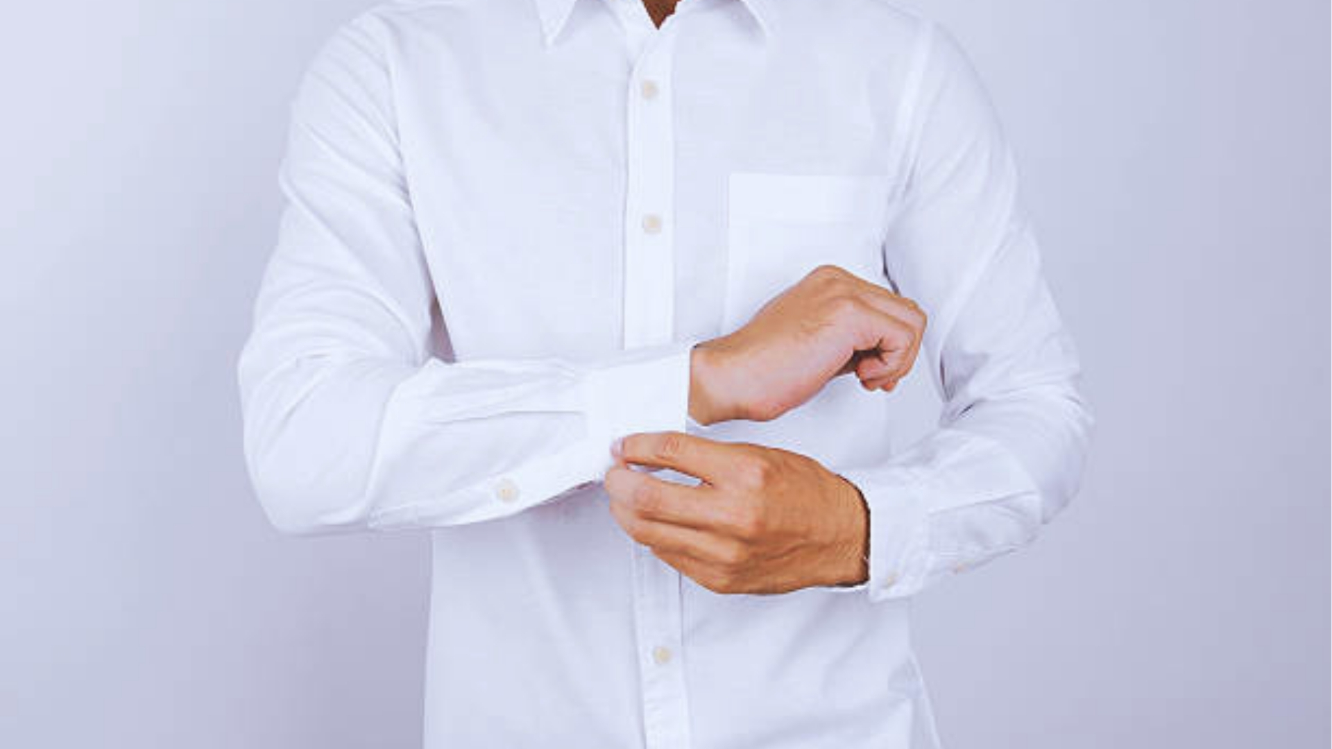 Close-up of man fastening cuff button on crisp white dress shirt with chest pocket against light gray background.
