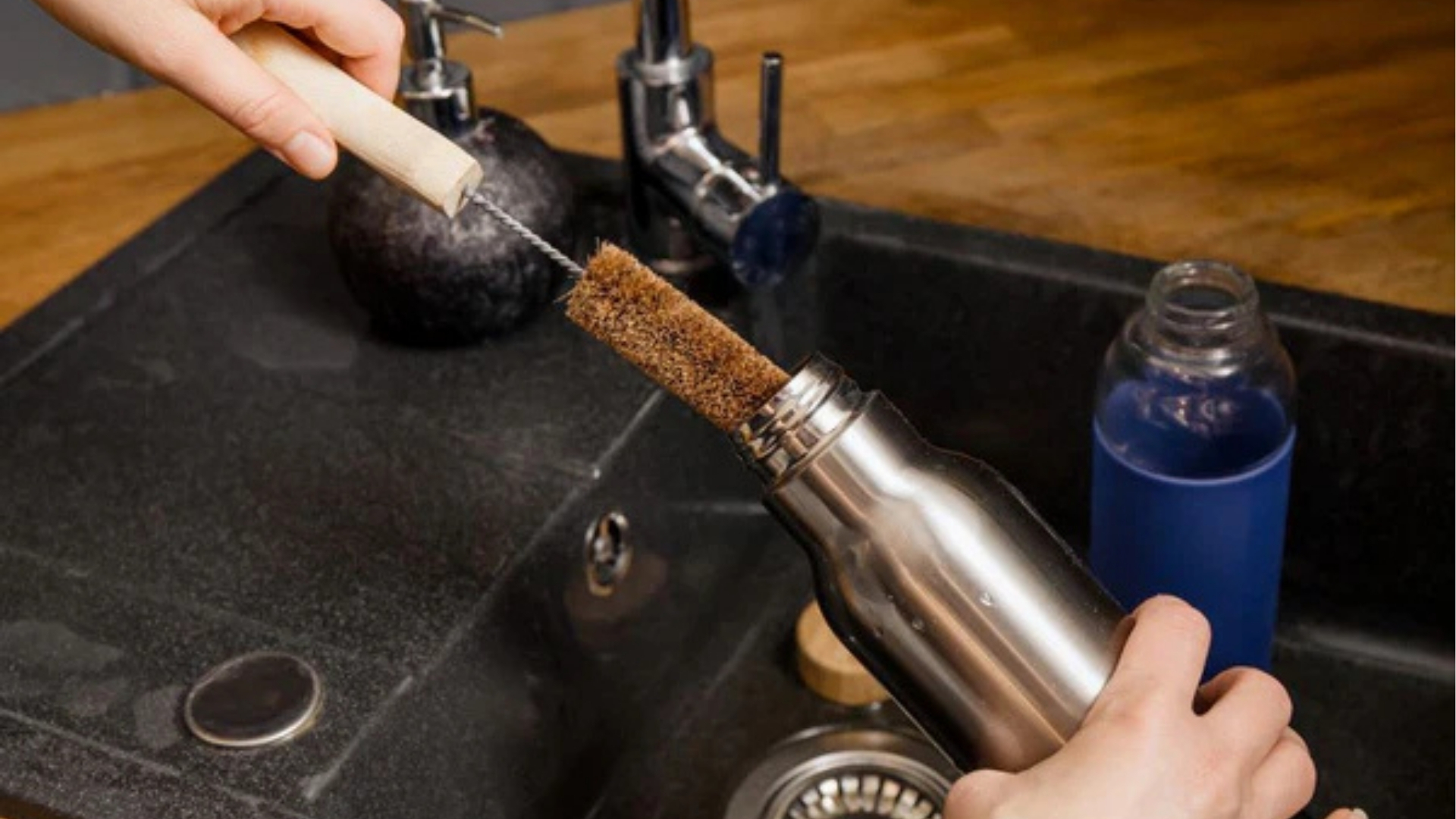 Person scrubbing inside stainless steel water bottle with long brown bristle brush over black kitchen sink