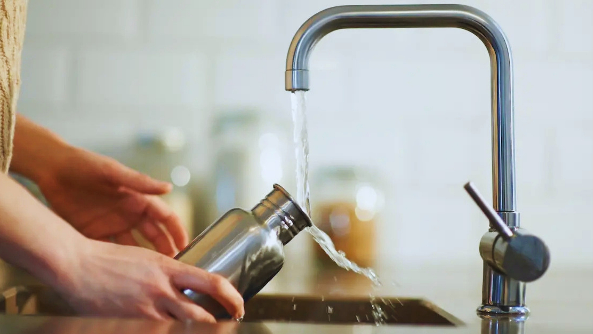 Person rinsing stainless steel water bottle under running tap from modern chrome faucet in bright kitchen with white tile backsplash.