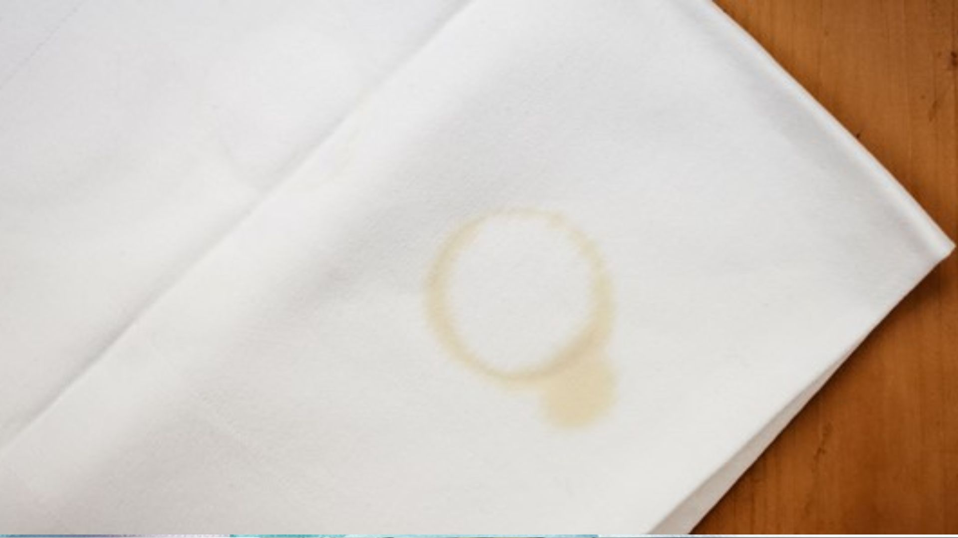 Top-down view of a folded white cloth napkin on a wooden table with a light brown circular oil ring stain and a small drip mark.