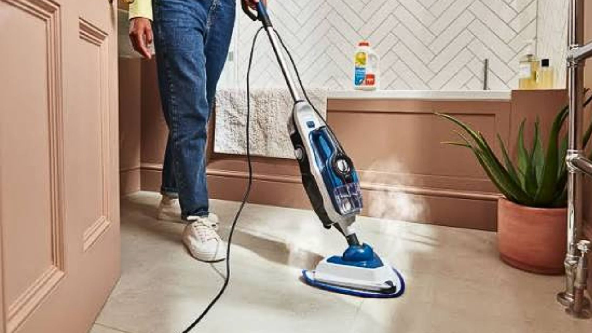Person pushing a blue and white steam mop across a tiled bathroom floor near a plant.
