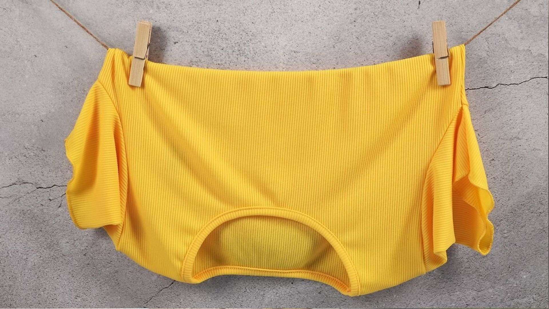 Bright yellow ribbed off-shoulder ruffled crop top hanging on a twine clothesline with two wooden clothespins against a gray concrete wall.