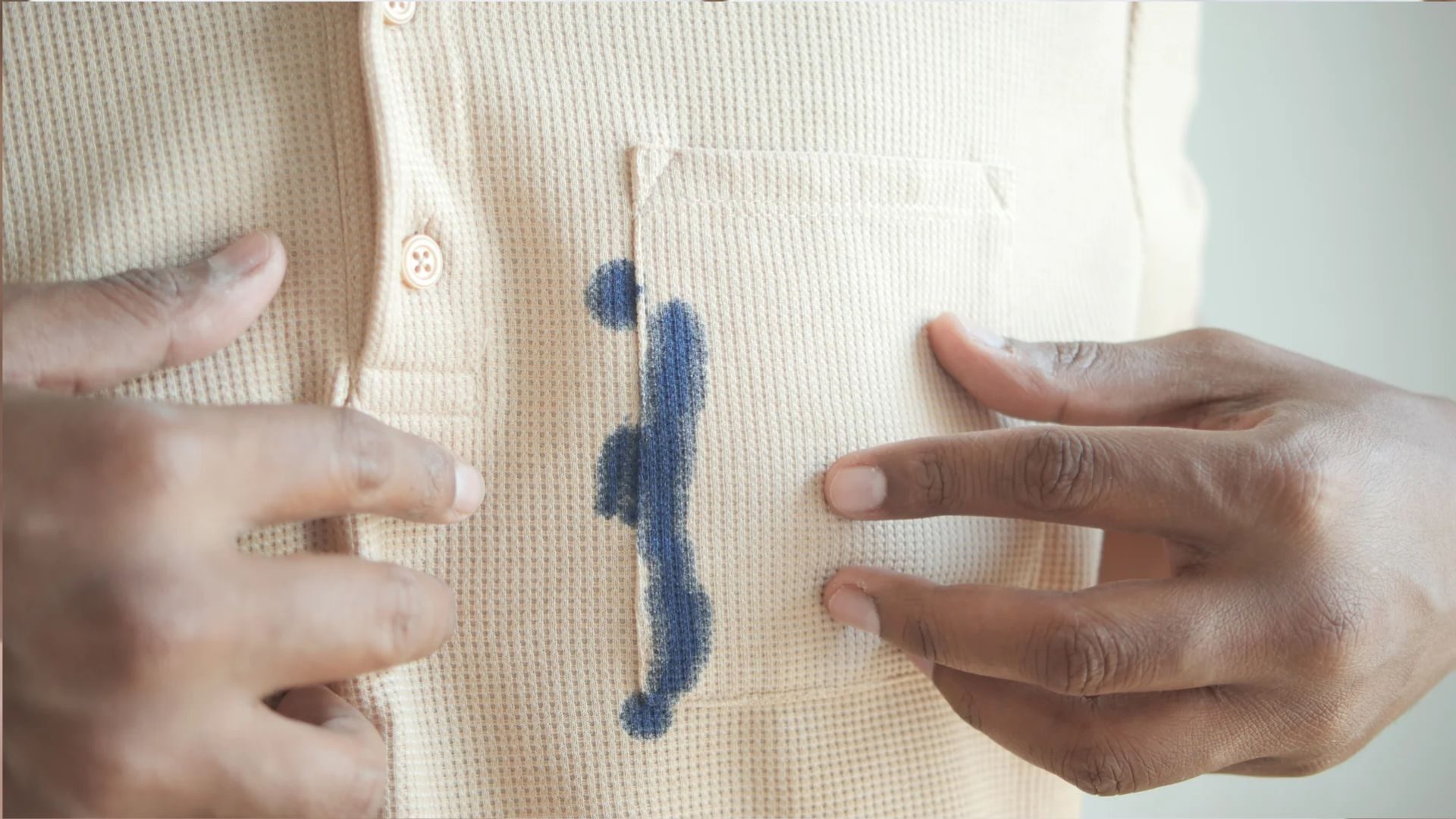 Close-up of hands holding a beige waffle-knit shirt with a prominent dark blue ink stain running vertically down the chest pocket area.
