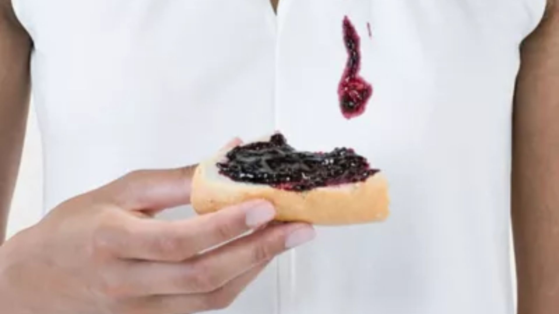 Person holding a slice of bread topped with dark berry jam, with a large purple-red jam stain dripped onto the front of their white shirt.