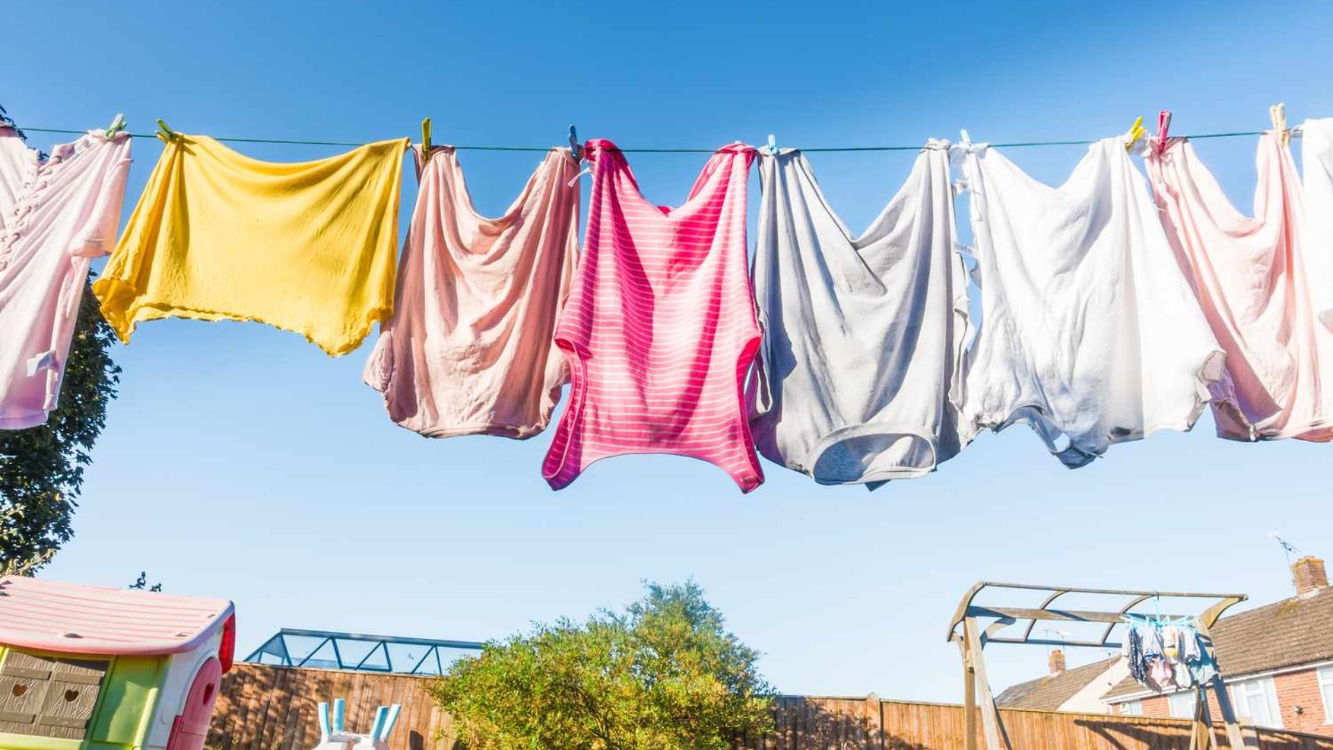 Colorful laundry hanging on outdoor clothesline under blue sky, with backyard, fence and playhouse below.
