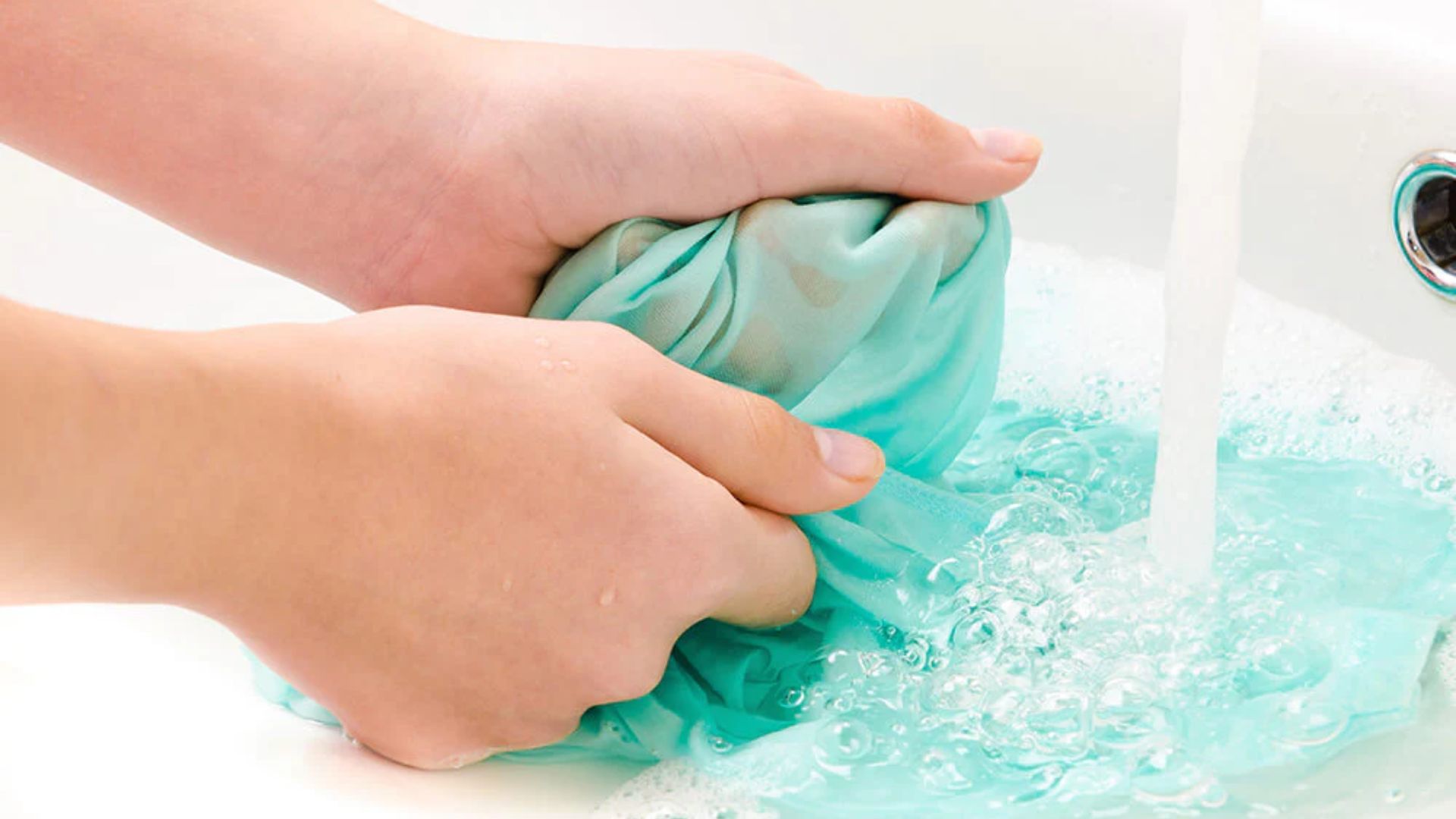 Hands wringing turquoise fabric under running water in sink with soap bubbles and suds.