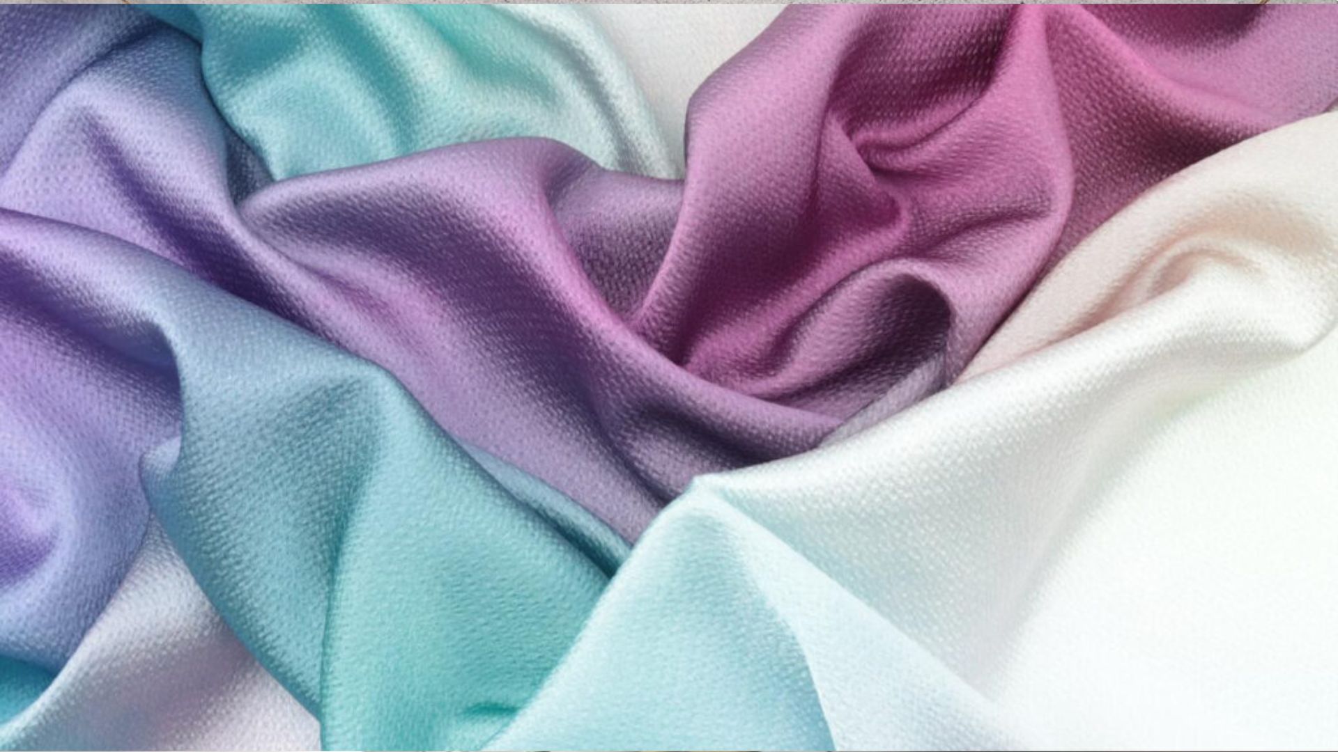 Close-up of soft shiny satin fabric in pastel lavender, turquoise, magenta and white, draped in gentle folds with textured surface detail.