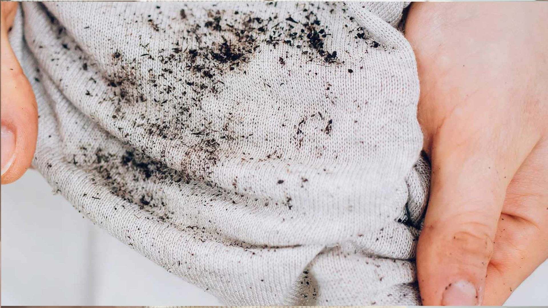 Close-up of hands holding a light gray fabric shirt covered with scattered dark black dirt and soil particles across the textured knit.
