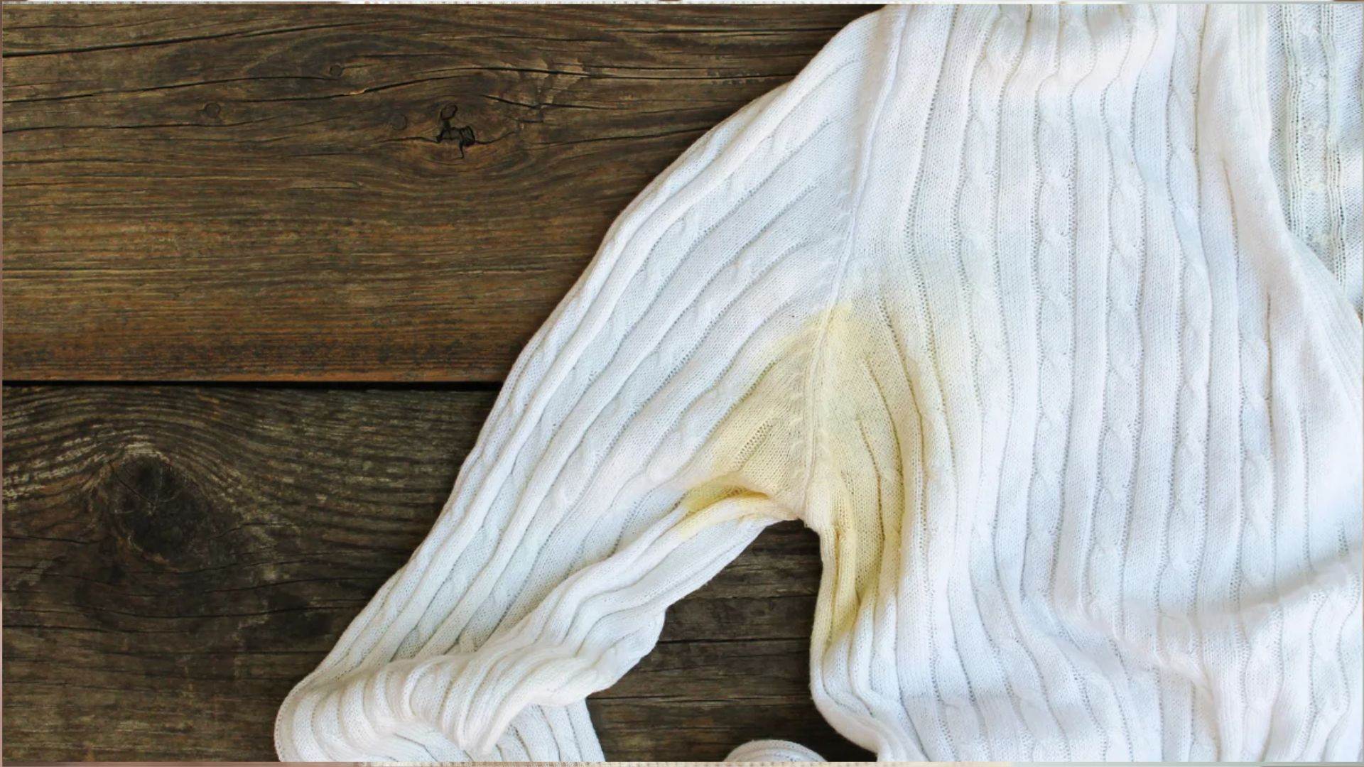 White cable-knit sweater sleeve on dark rustic wooden planks, showing a large yellowish deodorant or sweat stain around the underarm area.