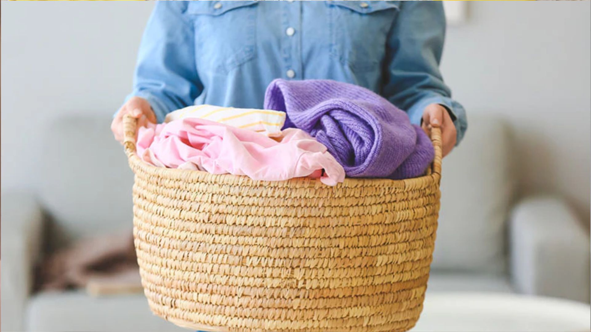 Person in blue denim shirt holding a woven straw laundry basket filled with colorful clothes including pink, purple and yellow garments.