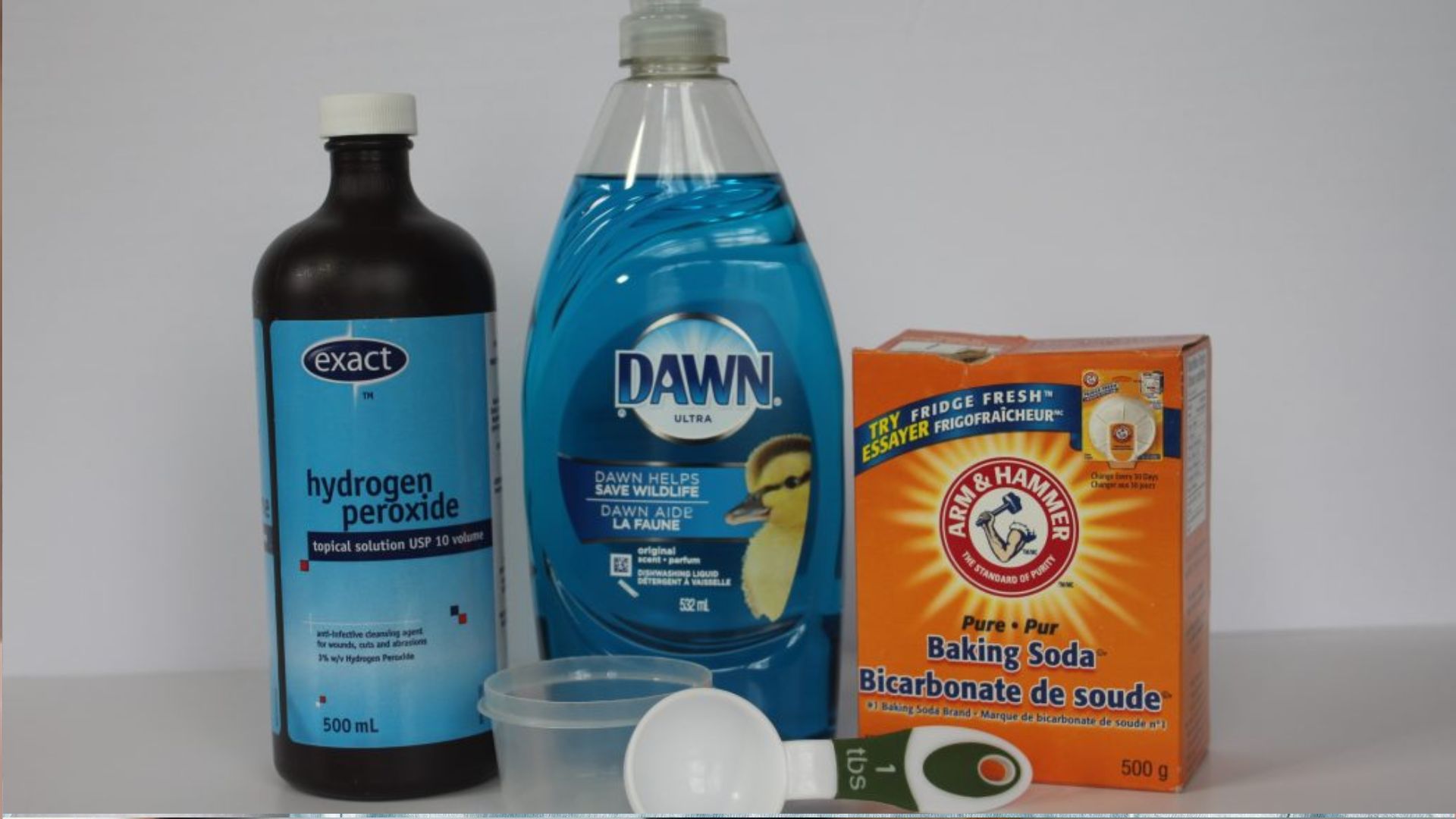 Three cleaning products on white: brown bottle of hydrogen peroxide, blue Dawn dish soap, and orange box of Arm & Hammer baking soda.